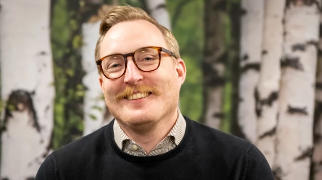 En man med glasögon och mustasch står framför en målning