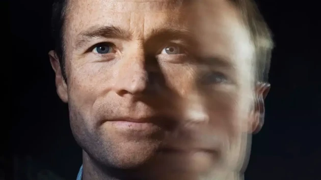 A man with short brown hair looking at the camera. The image is double exposed so it appears as though there is a blurry second image of the man to the right and slightly below the sharp image. 