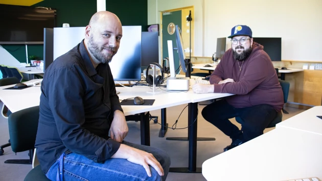 Två män sitter vid ett skrivbord och poserar framför kameran.