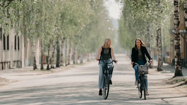 Two women cycling side-by-side on a birch-lined street leading to campus. 